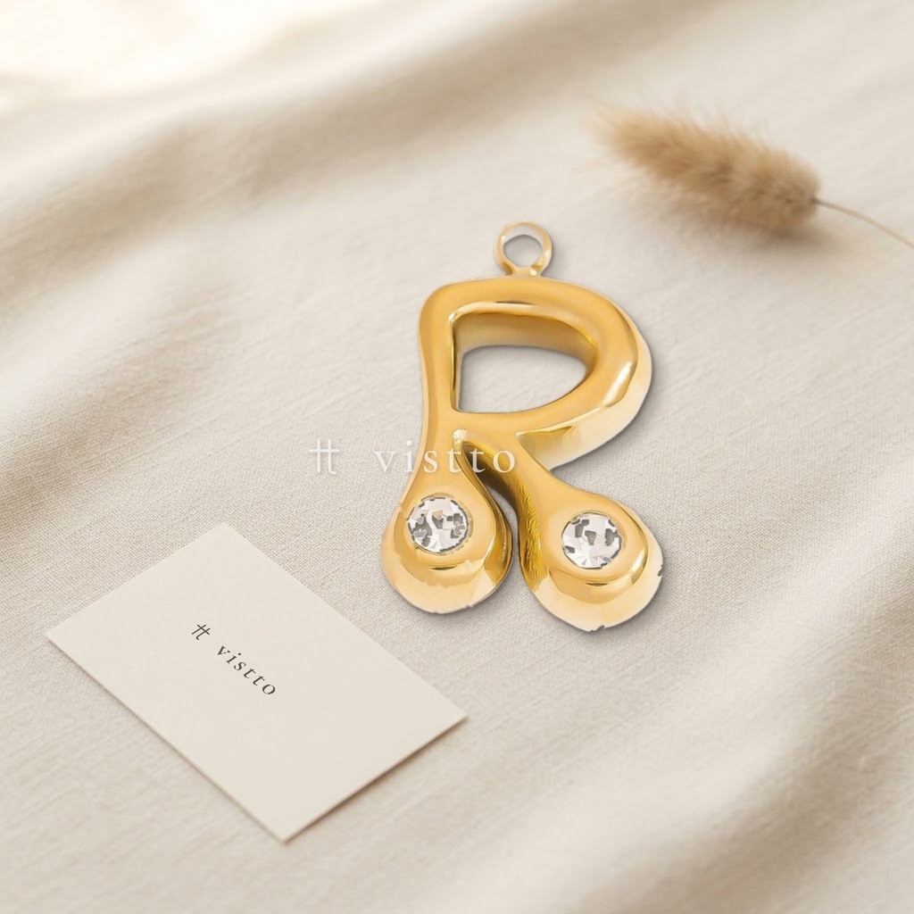 Dije dorado en forma de letra R de trazo abombado y liso, con circonias transparentes en los extremos y argolla superior